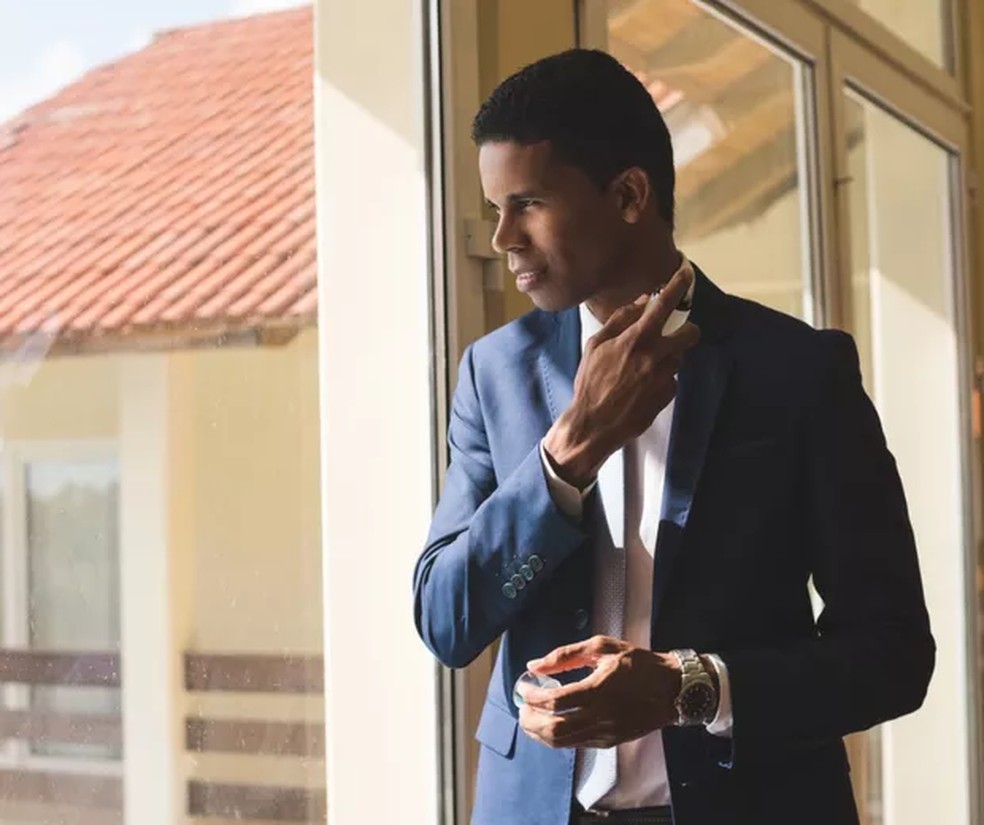 Homem negro de cabelos curtos usando um terno azul marinho passando perfume  Foto: Getty Images